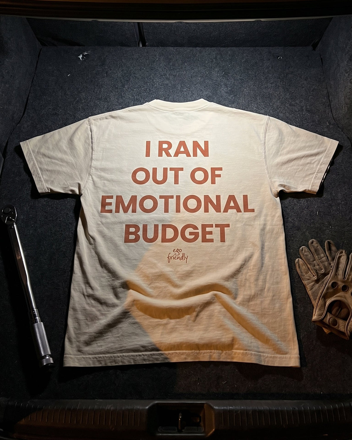 A light beige crewneck t-shirt with the phrase 'I RAN OUT OF EMOTIONAL BUDGET' printed in a terracotta orange font across the back, displayed inside a dark car trunk alongside a baseball glove and a tool.