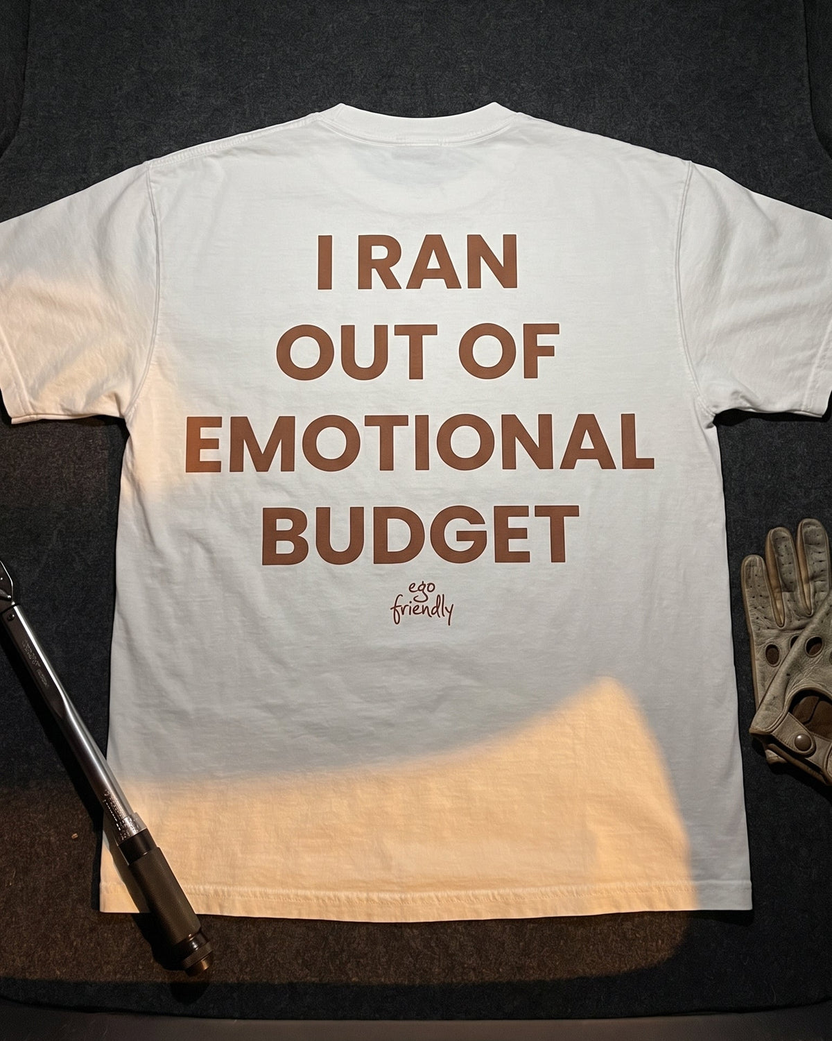 A white short-sleeve T-shirt laid flat on a dark surface, featuring the phrase 'I RAN OUT OF EMOTIONAL BUDGET' in large brown text across the back. A small script logo appears below the text.