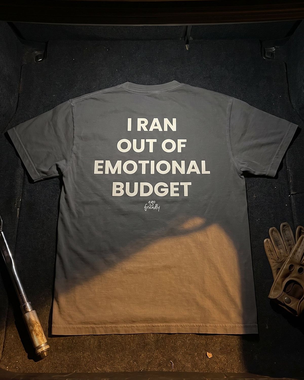 A dark gray t-shirt is laid flat inside an open car trunk, showing the back with white text that reads 'I RAN OUT OF EMOTIONAL BUDGET'. A baseball bat, leather gloves, and a bottle are placed around it as props.