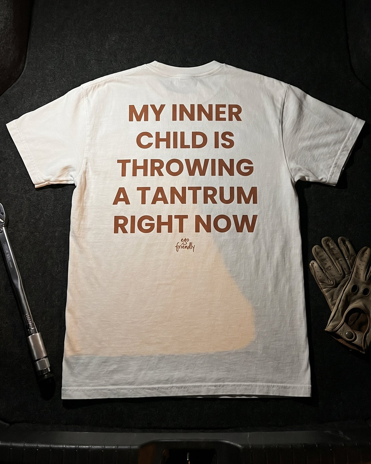A white graphic t-shirt is laid flat on a dark surface, showing the back side with the text 'MY INNER CHILD IS THROWING A TANTRUM RIGHT NOW' in brown. A small signature graphic is printed below the text.
