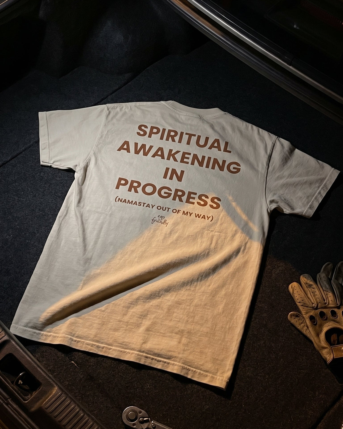 A light gray crewneck t-shirt with brown text reading 'SPIRITUAL AWAKENING IN PROGRESS (HAMASTAY OUT OF MY WAY)' laid flat in the trunk of a car, alongside a leather glove and a wrench.