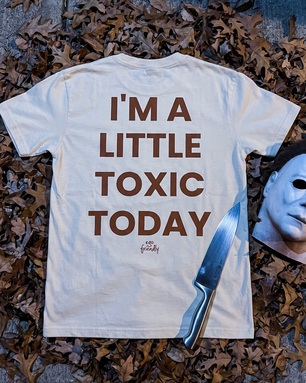 A white graphic t-shirt with brown text reading 'I'M A LITTLE TOXIC TODAY' is laid flat on a sidewalk covered with dry autumn leaves. A silver knife and a white mask are placed beside it.