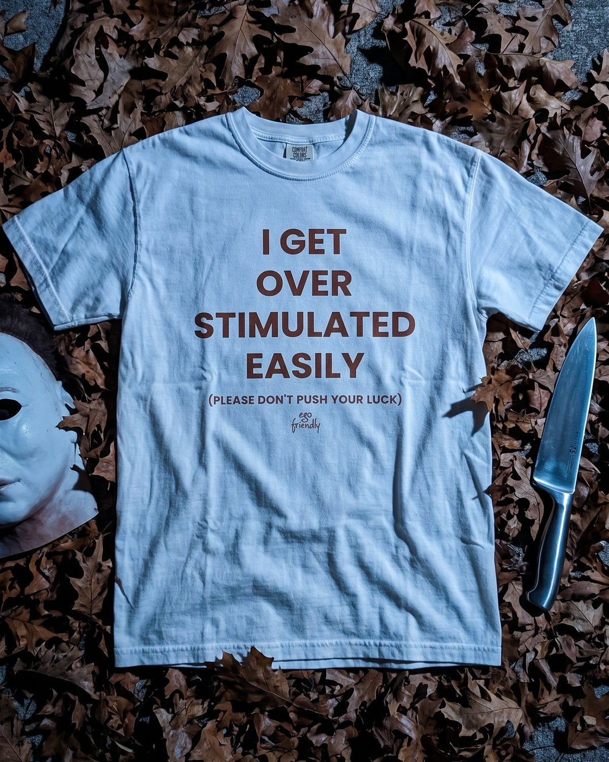A light blue graphic t-shirt with brown text reading 'I GET OVER STIMULATED EASILY' and a smaller line below, laid flat on a dark surface surrounded by scattered brown leaves, with a knife and a white mask prop nearby.