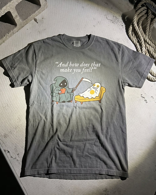 A dark gray graphic t-shirt laid flat on a concrete surface, featuring a cartoon character eating pizza with the text '"And how does that make you feel?"'.