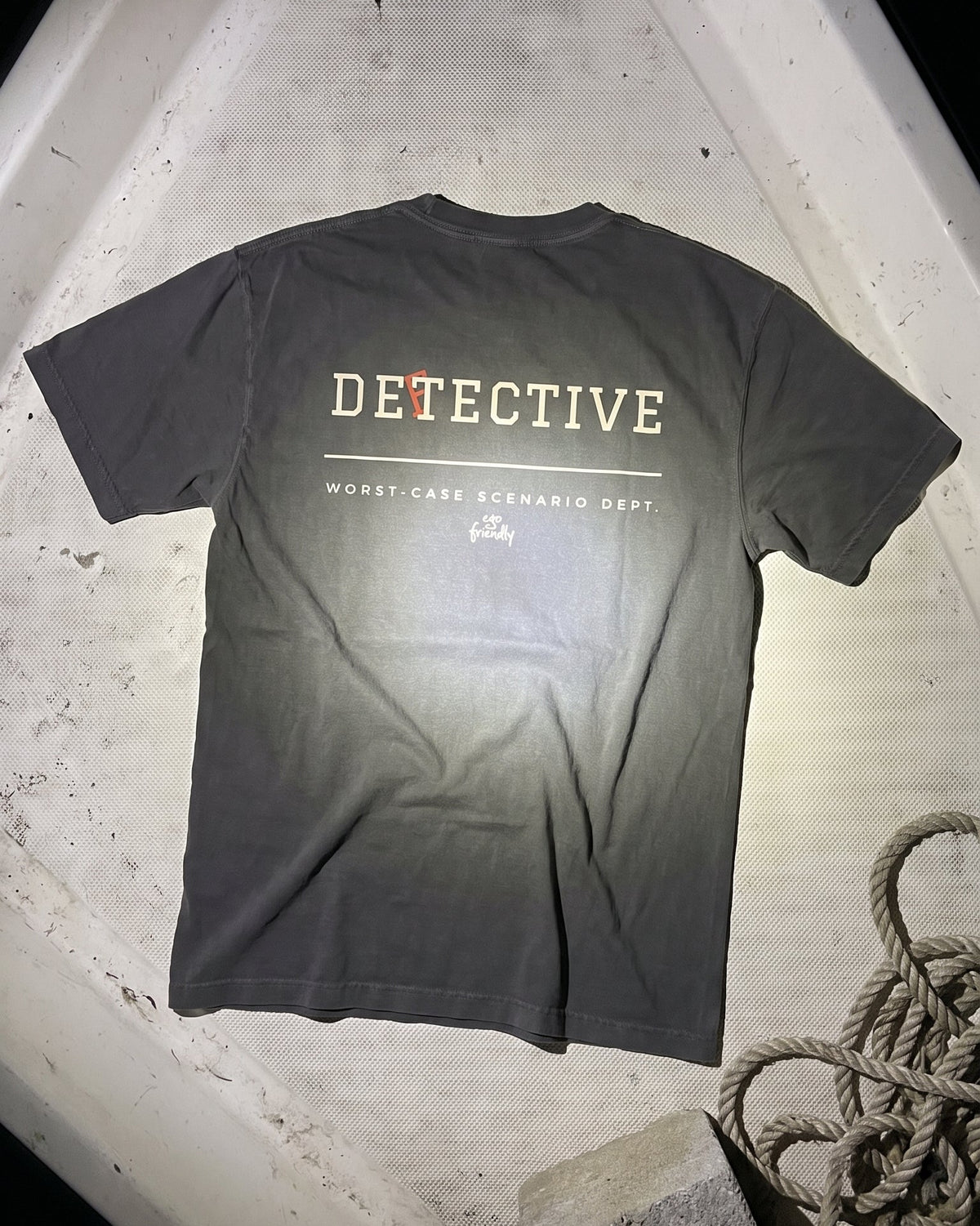 A dark gray t-shirt with 'DETECTIVE' and 'WORST-CASE SCENARIO DEPT.' printed on the back, laid flat on a weathered white surface with rope in the corner.