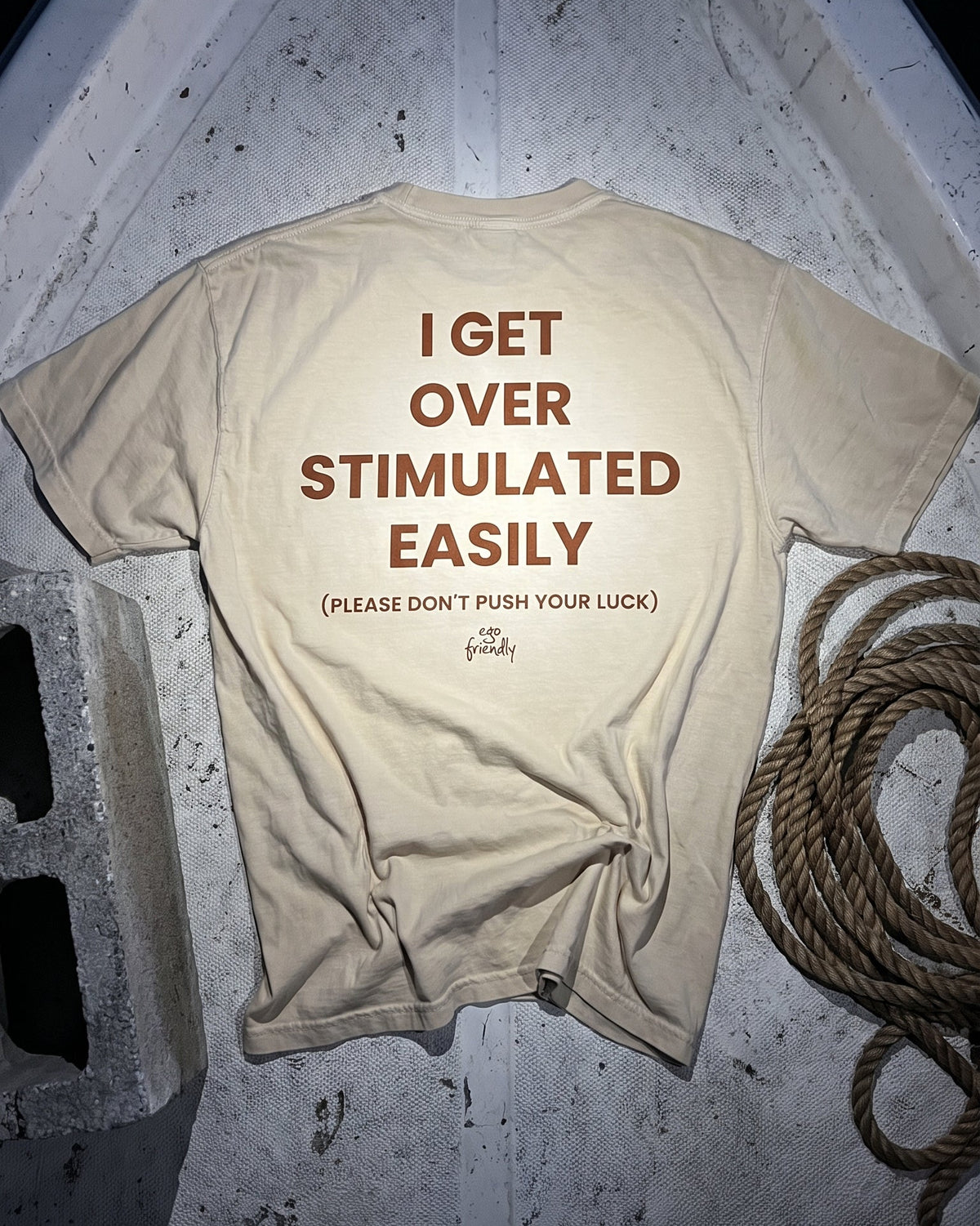 A beige graphic t-shirt laid flat on a textured gray surface, featuring brown text that reads 'I GET OVER STIMULATED EASILY (PLEASE DON'T PUSH YOUR LUCK)' with a small 'The Finalist' logo below.