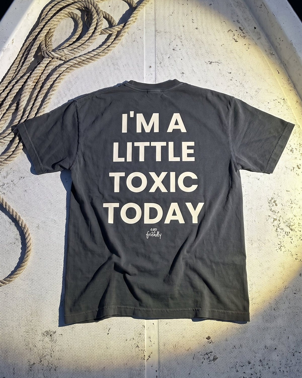 A dark gray t-shirt with the text 'I'M A LITTLE TOXIC TODAY' printed in white on the back, laid flat on the deck of a boat.