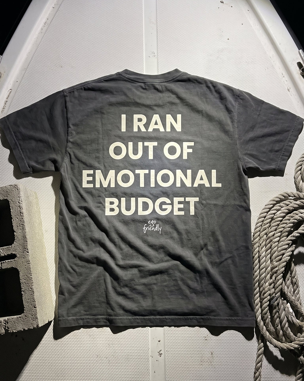 A dark gray crewneck t-shirt with the phrase 'I RAN OUT OF EMOTIONAL BUDGET' printed in bold white capital letters on the back, laid flat on a white boat deck next to a coiled rope.