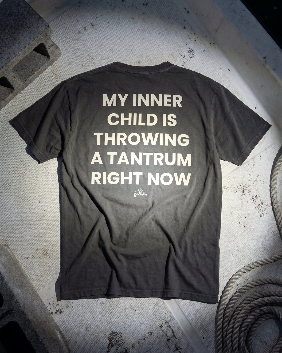 A dark charcoal gray t-shirt is laid flat on a concrete surface, showing the back with bold white text that reads 'MY INNER CHILD IS THROWING A TANTRUM RIGHT NOW'.