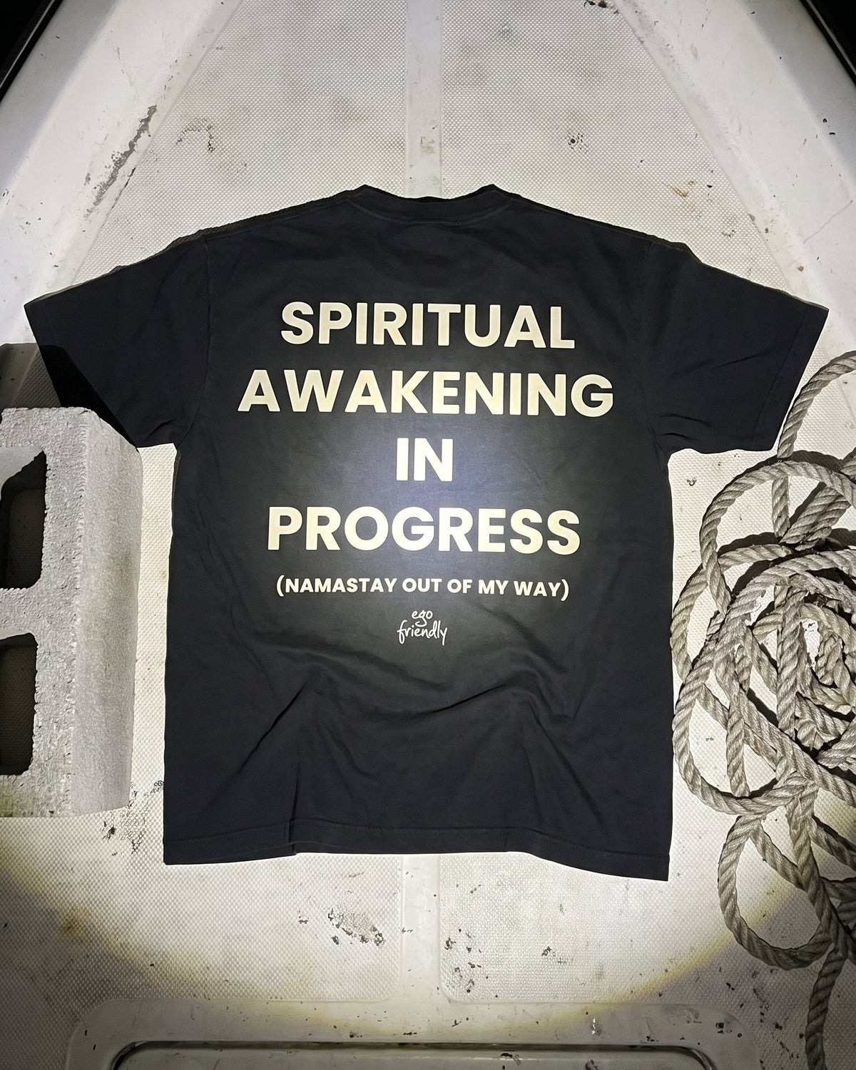A black t-shirt is laid flat on the deck of a boat under a spotlight, showing the back with bold cream text that reads 'SPIRITUAL AWAKENING IN PROGRESS (NAMASTAY OUT OF MY WAY)'.