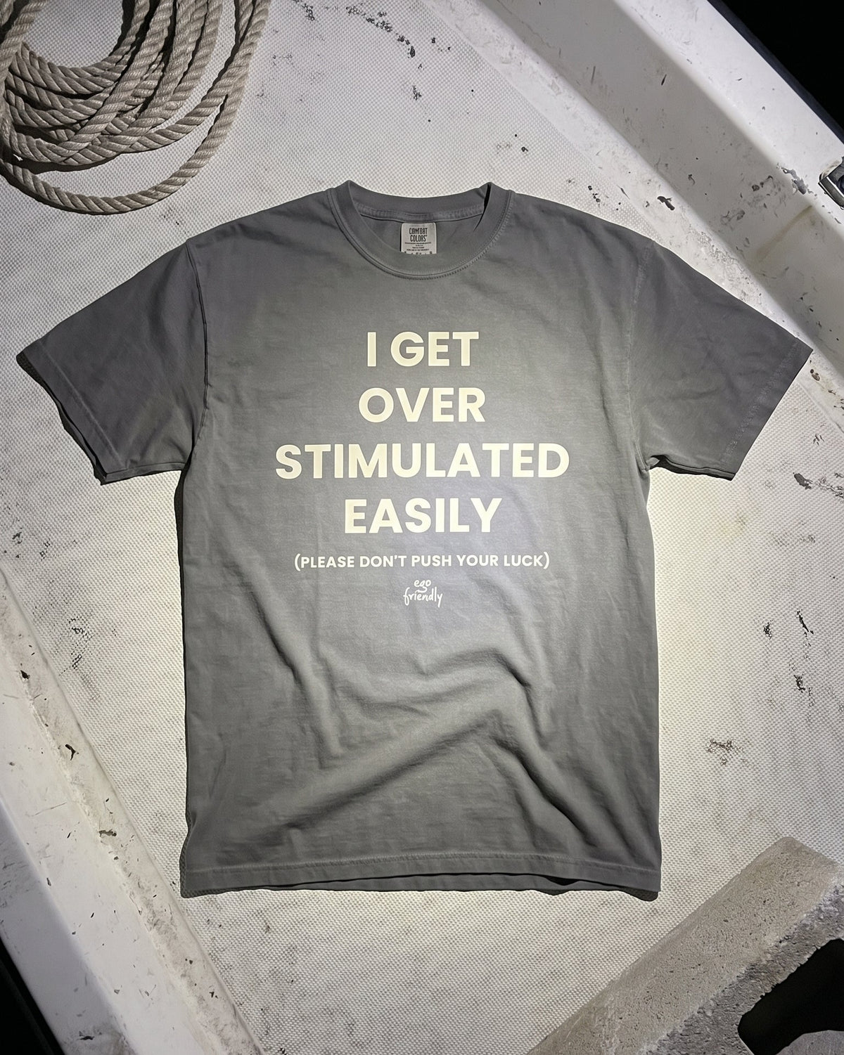 A gray short-sleeve t-shirt with cream-colored text reading 'I GET OVER STIMULATED EASILY' and a smaller line below, laid flat on a weathered white surface with a coiled rope in the top-left corner.