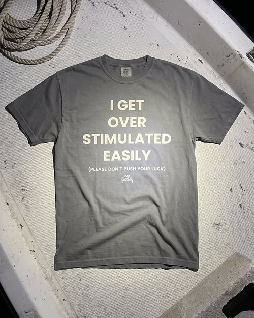 A gray short-sleeve t-shirt with cream-colored text reading 'I GET OVER STIMULATED EASILY' and a smaller line below, laid flat on a weathered white surface with a coiled rope in the top-left corner.