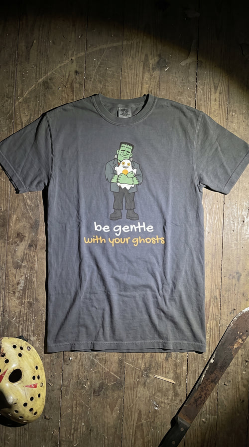 A dark gray graphic t-shirt featuring a cartoon of Frankenstein holding a ghost, with the text 'be gentle with your ghosts' printed below. The shirt is laid flat on a rustic wooden surface next to a hockey mask and a machete prop.