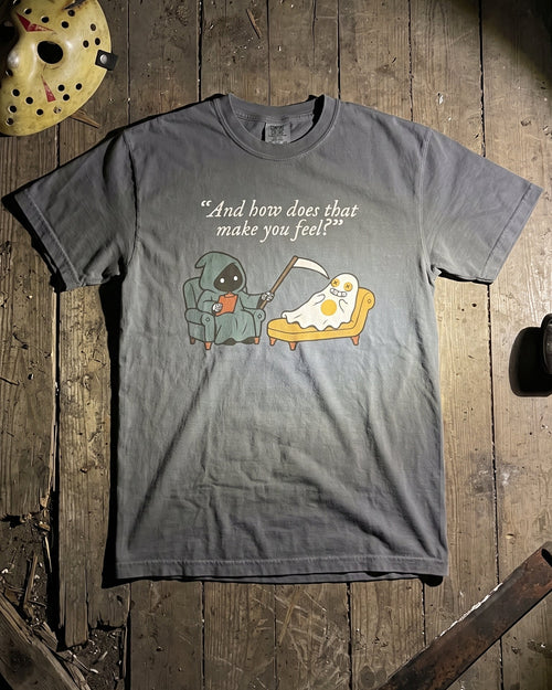 A gray graphic t-shirt featuring a cartoon of a character eating pizza and the text '"And how does that make you feel?"', laid flat on a rustic wooden plank background with a hockey mask prop in the corner.
