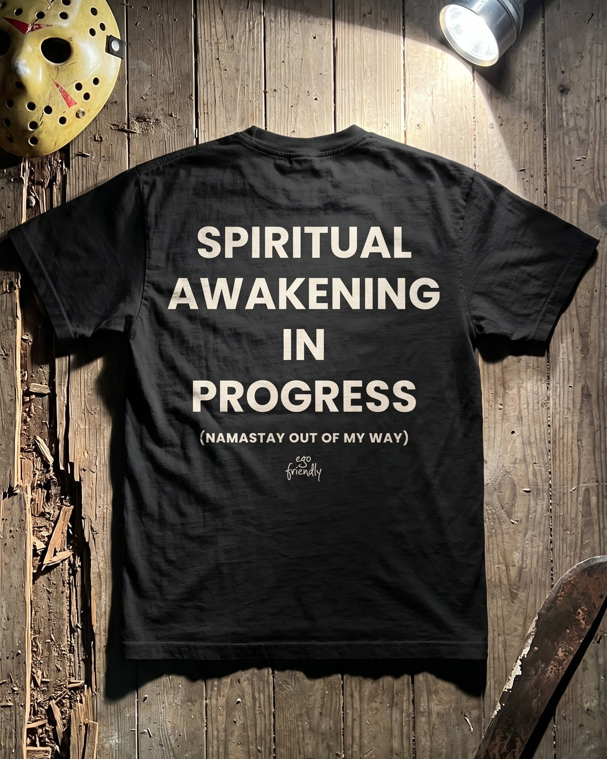 A black graphic t-shirt is laid flat on a rustic wooden background, illuminated by a flashlight. The back of the shirt features the text 'SPIRITUAL AWAKENING IN PROGRESS (NAMASTAY OUT OF MY WAY)' in white.