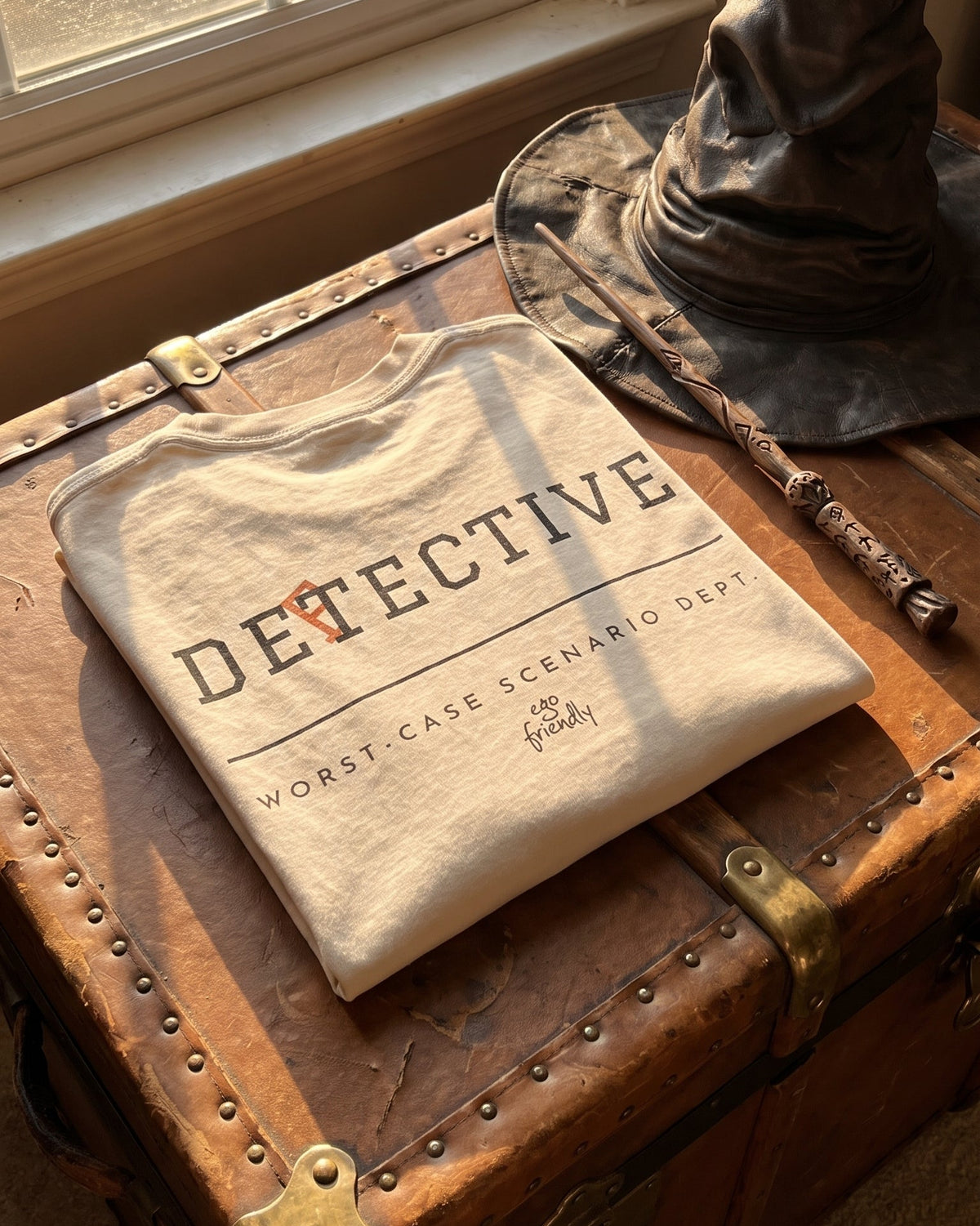 A folded beige graphic T-shirt with the words 'DETECTIVE' and 'WORST-CASE SCENARIO DEPT.' lies on a rustic brown leather trunk.
