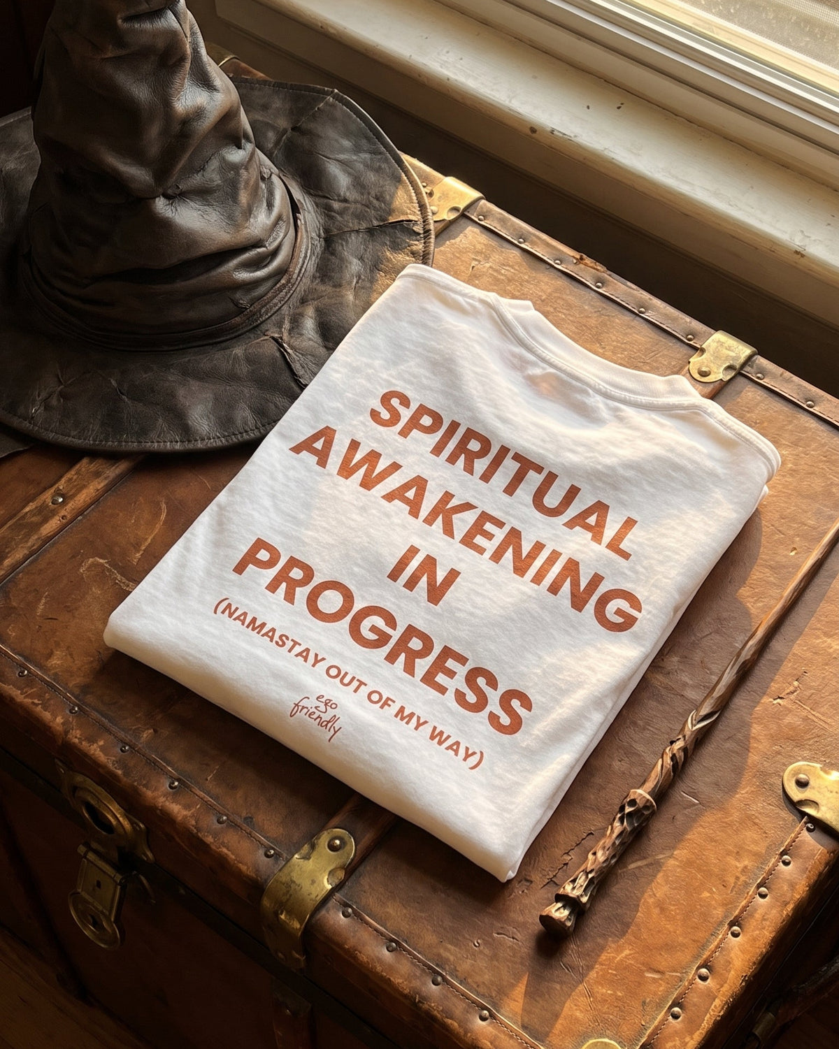 A folded white graphic T-shirt with orange-red text reading 'SPIRITUAL AWAKENING IN PROGRESS (WAMASTAY OUT OF MY WAY)' is displayed on a rustic brown wooden trunk beside a dark pointed hat and wand, near a window.