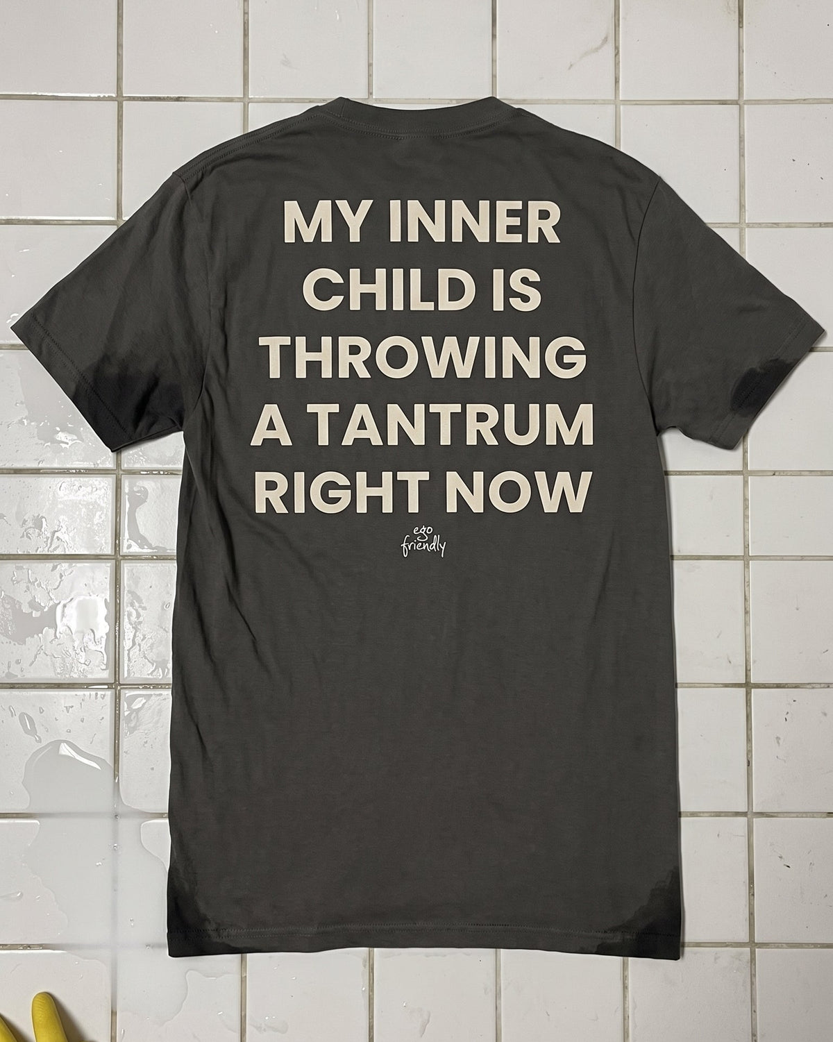 A dark gray t-shirt with bold cream-colored text on the back reading 'MY INNER CHILD IS THROWING A TANTRUM RIGHT NOW' and a small 'the giddy' logo underneath, laid flat on a white tiled wall with a yellow glove in the corner.