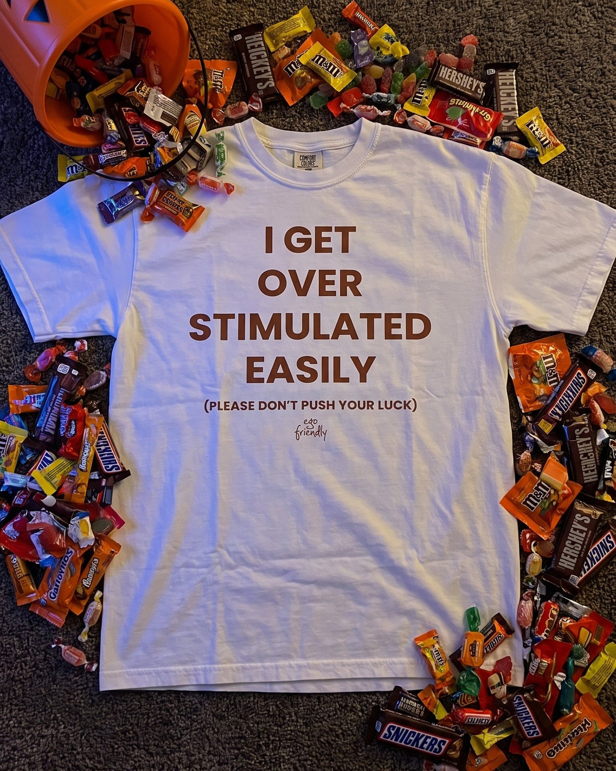 A white graphic t-shirt with brown text reading 'I GET OVER STIMULATED EASILY' is laid flat on a dark carpet, surrounded by an explosion of candy and an orange jack-o'-lantern bucket overflowing with treats.