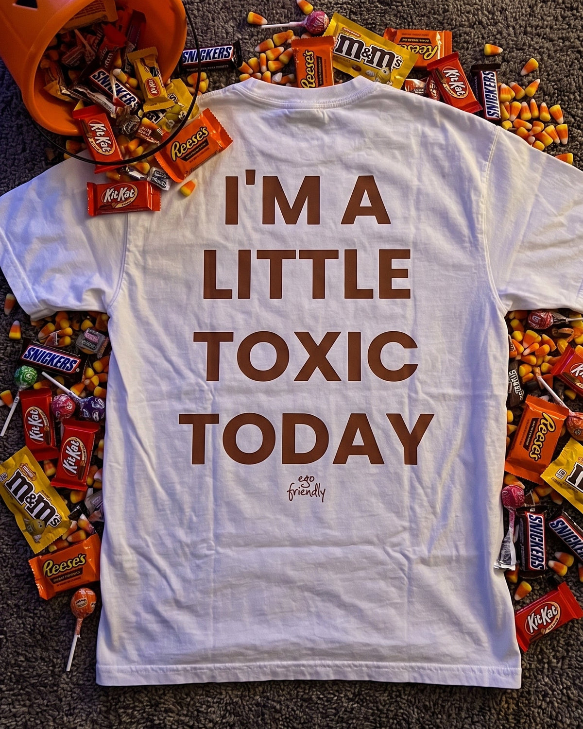 A white graphic T-shirt laid flat on a dark carpet, surrounded by Halloween candy and an orange bucket. The shirt has large brown text reading 'I'M A LITTLE TOXIC TODAY' with a small 'The Toxic Society' logo underneath.