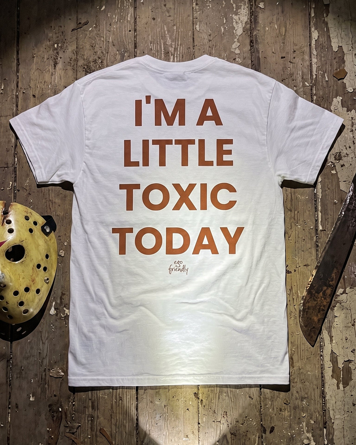 A white short-sleeve t-shirt laid flat on a rustic wooden surface, showing the back printed with the phrase 'I'M A LITTLE TOXIC TODAY' in orange-brown lettering. A hockey mask, a machete, and a flashlight are placed around it as styled props.