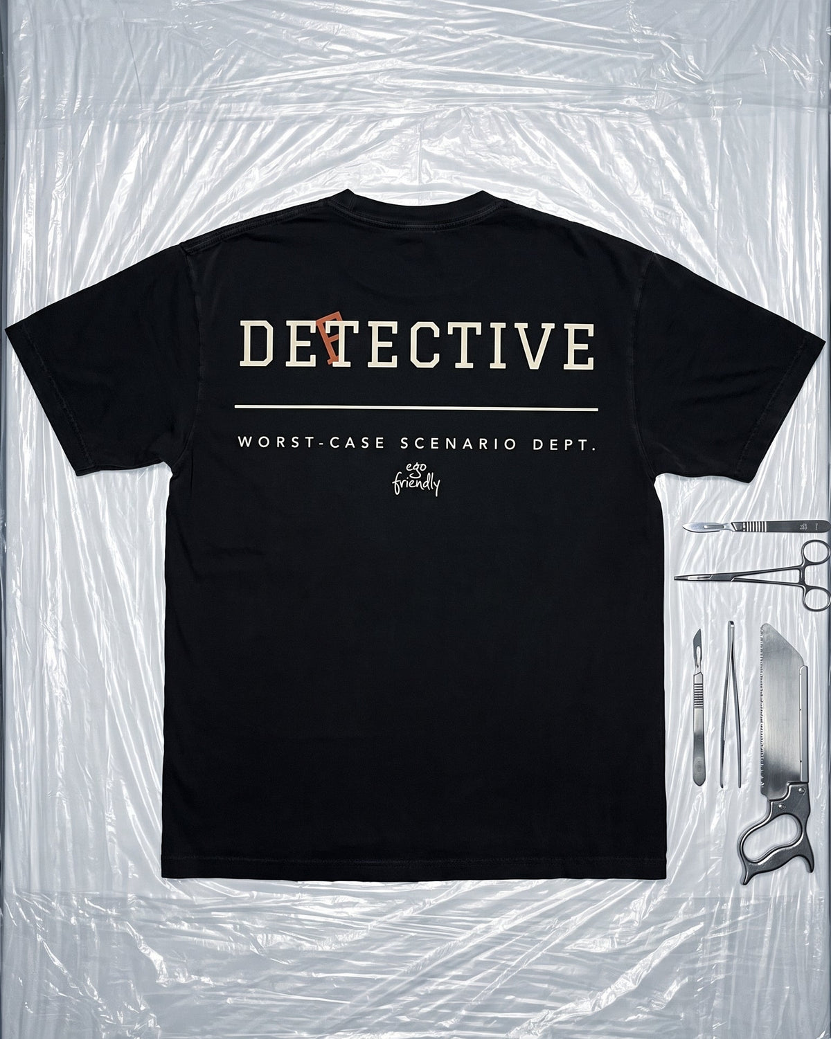 A black short-sleeve t-shirt with 'DETECTIVE' and 'WORST-CASE SCENARIO DEPT.' printed on the back, laid flat on a crinkled clear plastic sheet with surgical tools arranged to the right.