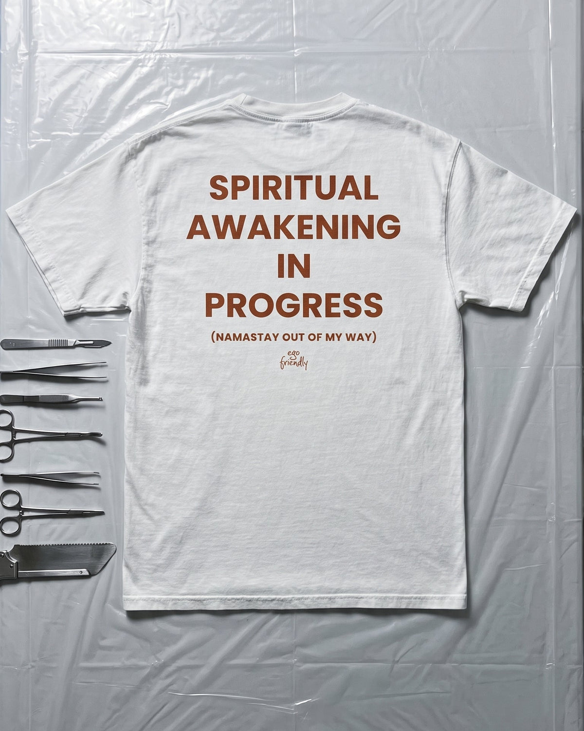 A white short-sleeve T-shirt laid flat on a clear plastic-covered surface, with bold brown text reading 'SPIRITUAL AWAKENING IN PROGRESS' and smaller text below. Surgical scissors are placed along the left edge as styling props.