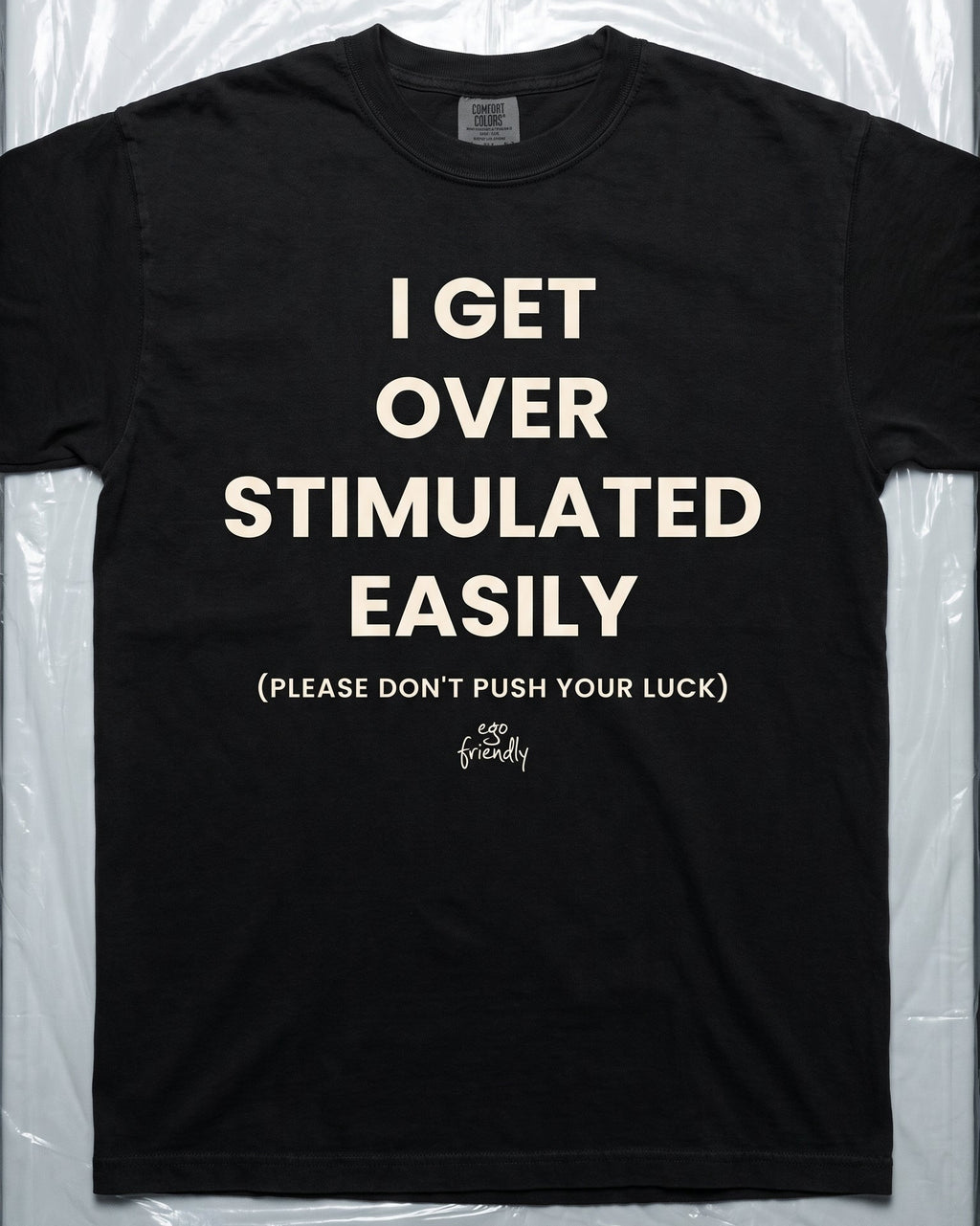A black graphic t-shirt with the text 'I GET OVER STIMULATED EASILY (PLEASE DON'T PUSH YOUR LUCK)' in cream lettering, laid flat on a white background with medical tools like forceps and scissors above it.