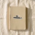 The Wordmark kiss-cut vinyl sticker applied to the oat cover of a plain notebook on cream linen in warm window light. It reads EGO over FRIENDLY in heavy black type, with a fried egg in place of the O in EGO.